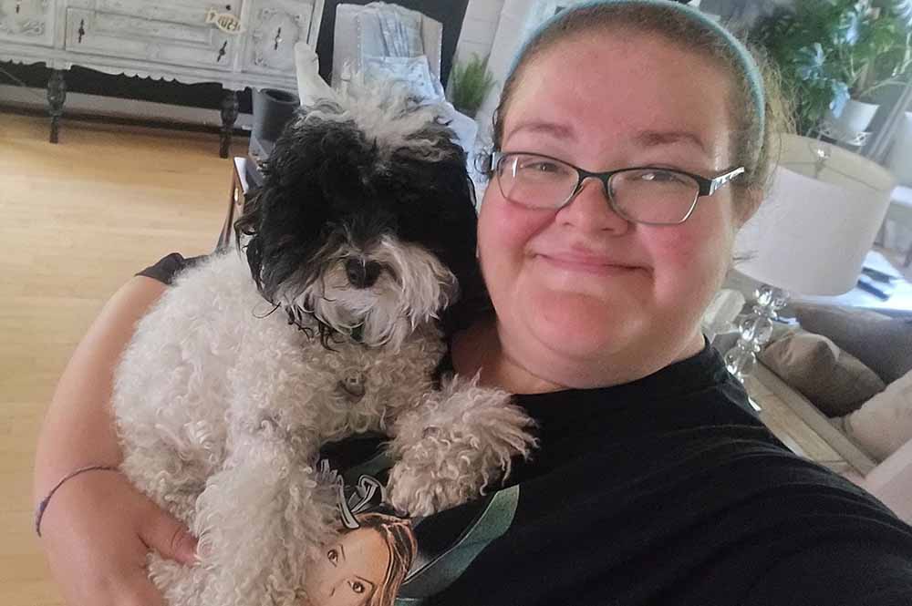 A woman wearing glasses gently holds a small white dog in her arms, smiling warmly at the camera